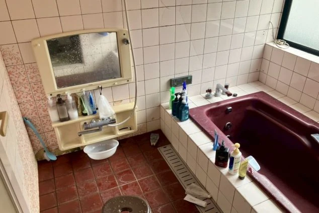 シェア寮の浴室。赤茶色の浴槽とタイル、壁には棚付きの鏡と多くのシャンプー類が置かれている。