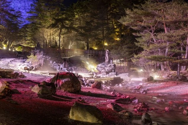 草津温泉の湯畑を背に、水玉模様の服を着た女性が後ろ姿で湯けむりを眺めている画像。勢いよく流れ落ちるお湯と緑の岩肌が特徴的。リゾートバイトの休日に温泉観光を楽しむイメージ。