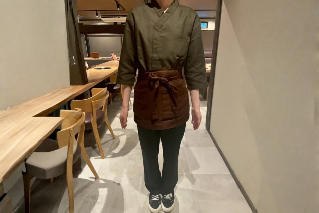 オリーブグリーン(またはカーキ色)の上着に、茶色の前掛け(エプロン)、黒いズボンとスニーカーを着用した、飲食店のスタッフと思われる人物。