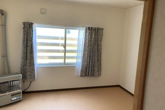 窓に花柄のカーテンがかかった、何も置かれていない部屋。床は木目調で、窓の横にはファンヒーターがある。