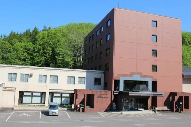 青い空と緑の森を背景に、赤茶色の外壁と白い横長の建物が組み合わさったホテル外観。