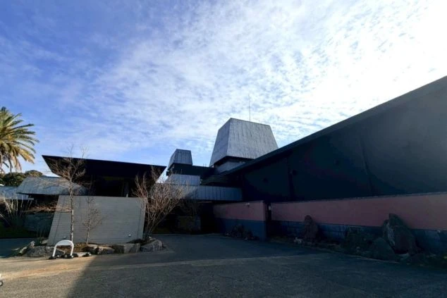 エントランスと建物外観。灰色のピラミッド型の屋根と、ベージュ色の壁、そして黒っぽい車寄せの庇（ひさし）が特徴的な建物。敷地内にはヤシの木が植えられている。