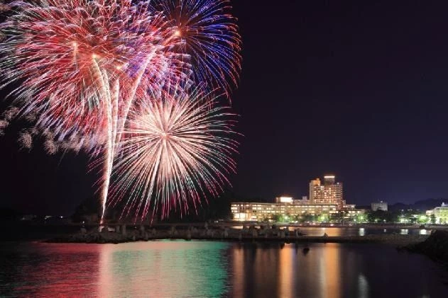 夜空に大きく打ち上がる色鮮やかな花火。水面に花火の光が反射し、遠くには明かりの灯った建物が見える。