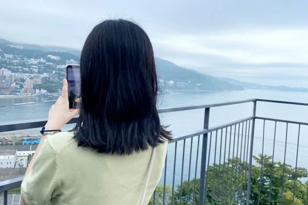 熱海の海岸沿いの展望台で、女性がスマートフォンで景色を撮影している後ろ姿の画像です。遠くには海と街並みが広がり、リゾートバイトの休日に絶景を楽しむ様子が伝わります。
