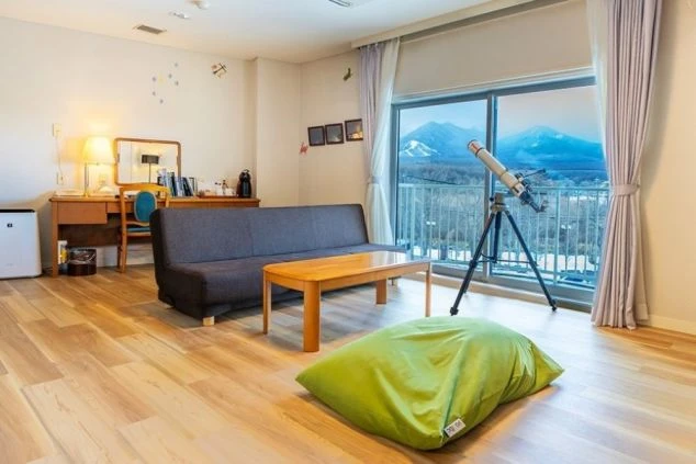 窓の外に雪をかぶった山々が見える、ホテルの客室の画像。望遠鏡が置かれており、自然をゆっくりと堪能できるリゾートの雰囲気を伝えている。