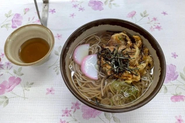 リゾートバイトのまかない画像。大きなかき揚げ、紅白のかまぼこ、刻み海苔がのった温かい天ぷらそば。