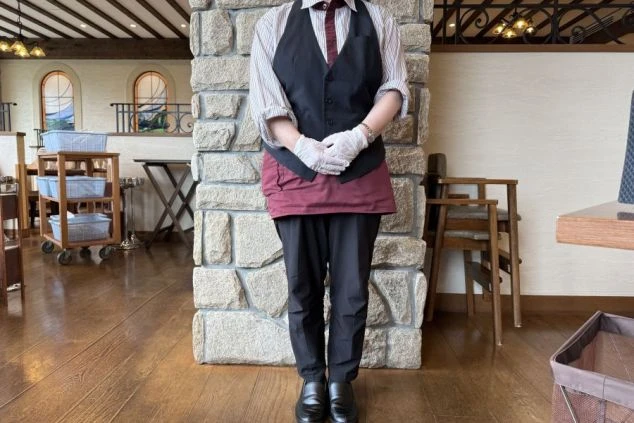 リゾートバイトのレストランスタッフ女性の制服画像。黒いベストと赤茶色のエプロン、ストライプのシャツの組み合わせで、清潔感があり動きやすいホールスタッフの服装。