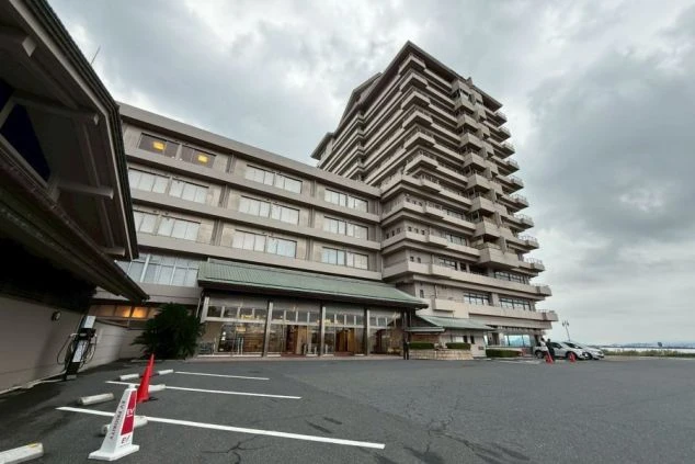 琵琶湖グランドホテルの正面外観。低層のロビー棟と、段々になったバルコニーが特徴的な高層棟。手前はアスファルトの駐車場で、奥には琵琶湖が見える。