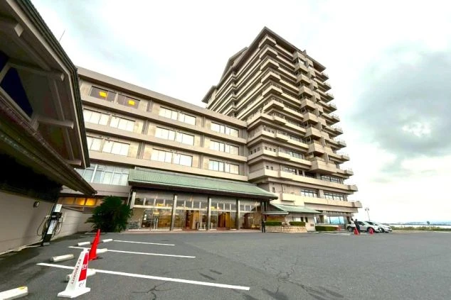 琵琶湖グランドホテルの正面外観。低層のロビー棟と、段々になったバルコニーが特徴的な高層棟。手前はアスファルトの駐車場で、奥には琵琶湖が見える。