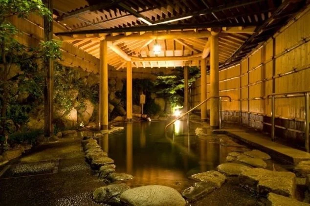 湯田温泉にあるホテルの露天風呂の画像。木製の屋根と竹垣に囲まれた湯舟が、夜の帳の中で幻想的にライトアップされている。リゾートバイトの勤務後にリラックスできる温泉施設。