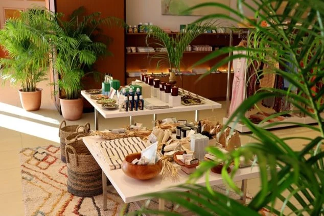 リゾートホテルの土産物店またはブティックの店内。観葉植物が豊富に置かれ、棚やテーブルの上には地元の手工芸品、アメニティ、衣類などが並べられている。