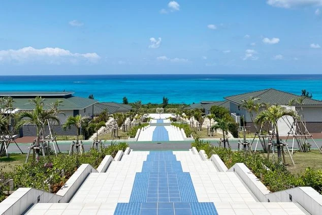 ターコイズブルーの海に向かって真っ直ぐに伸びる、白いタイルと青いモザイクで装飾された階段と通路。両脇にはリゾート風の低層の宿泊棟やヤシの木が並び、リゾート地の開放的な風景。