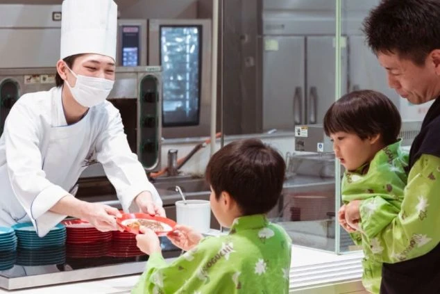 ホテルのビュッフェカウンターで、白いコック服とマスクを着けたシェフが、子供（浴衣姿）に料理の入った赤い皿を手渡している。隣には父親らしき男性と、もう一人の子供が立っている。