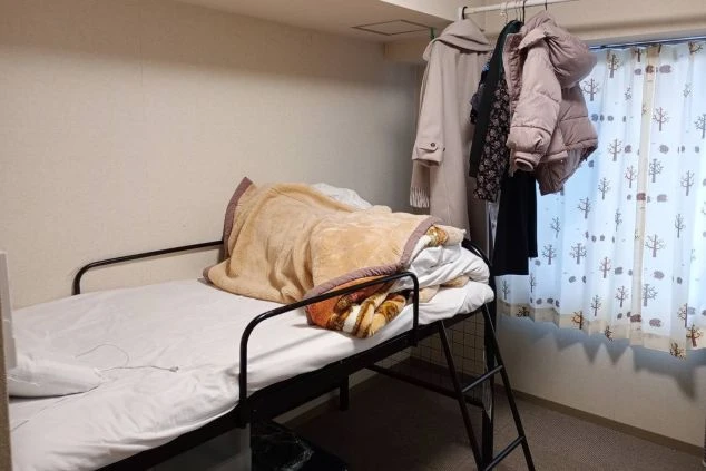 寮の居室。ベッドと窓、ハンガーラックに服がかかっている。簡素ながらも生活に必要なものが揃っている。