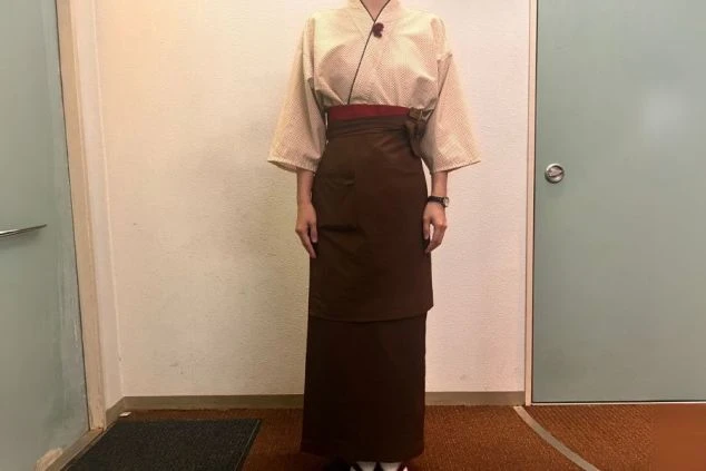 着物風の上着に濃い茶色の前掛けを着用した女性スタッフの画像。足元は草履を履いている。旅館や和食レストランでのリゾートバイトの仕事内容の一つである、ホテルサービススタッフの制服画像。