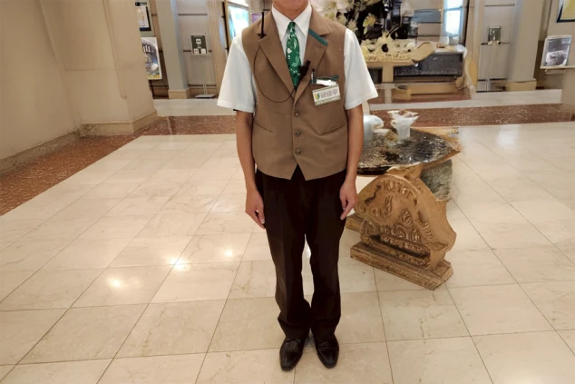 フロントスタッフの制服を着た男性。白いシャツに茶色いベストと緑色のネクタイを着用している。