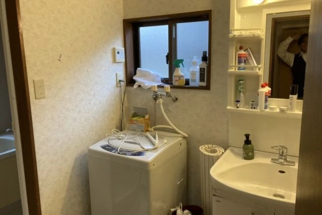 洗面台と洗濯機が並ぶ寮の洗面脱衣所の画像。窓から光が差し込み、洗剤や備品が整理されている。リゾートバイト中の身だしなみや洗濯に便利な設備。