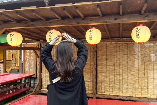 「おかげ横丁」と書かれた提灯が吊るされた軒下で、女性が後ろ姿で両手でハートマークを作っている画像。観光を楽しむ様子が伝わる。