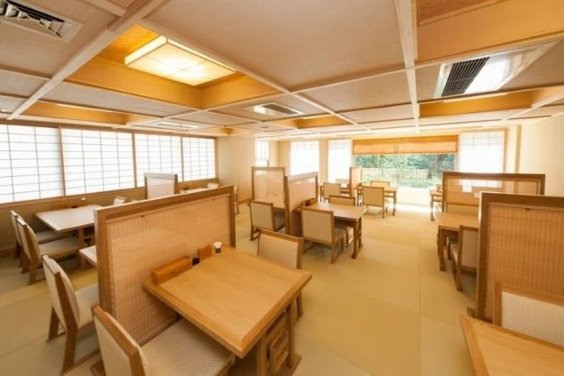 木と障子で構成された和風の広間レストランの画像。座敷席やテーブル席が並び、窓からは緑が見える。和食を提供する旅館やホテルのレストランのイメージ。