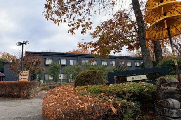 バリ風の黄色い傘と石像が置かれた、アンダの森 伊豆いっぺき湖の入口。奥にはシックな黒い外観の宿泊棟が見えます。