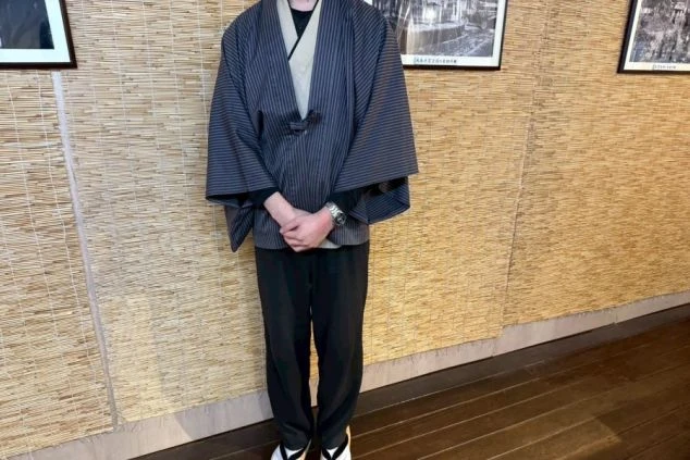 濃いグレーの縦縞の羽織のような上着に黒いパンツを着用し、足元は白足袋と草履。