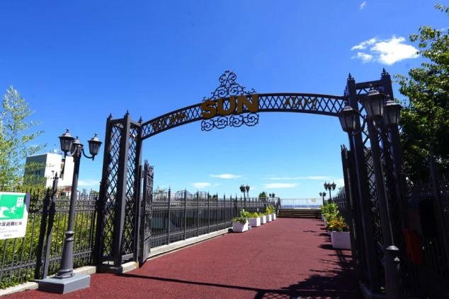 リゾートバイトの勤務先の外観を表現するイメージ画像。青空の下に「SUN」という文字が飾られたアイアンゲートがあり、その先には海へと続く遊歩道が広がっている。