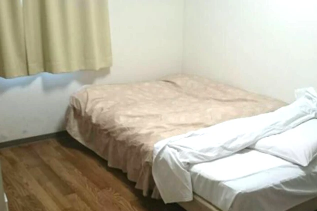 白い壁と木目調の床が特徴的な、寮の個室。シンプルなベッドが配置され、窓には淡いベージュのカーテンがかかっています。