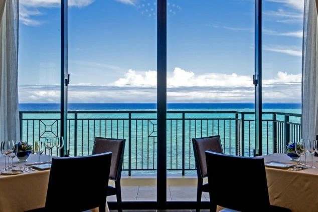 窓の外に広がるエメラルドグリーンの海と青い空が美しい、オーシャンビューのリゾートホテルのレストラン内部。リゾートバイトで絶景のレストランで接客や配膳の仕事ができることを示す魅力的な画像。