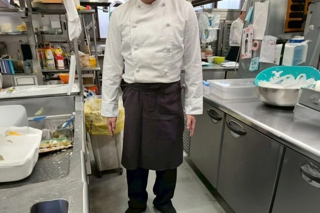 調理場の厨房に立つ男性スタッフの画像。白いコックコートと黒いズボン、黒いエプロンを着用している。リゾートバイトの仕事内容の一つである、調理補助スタッフの制服の画像。