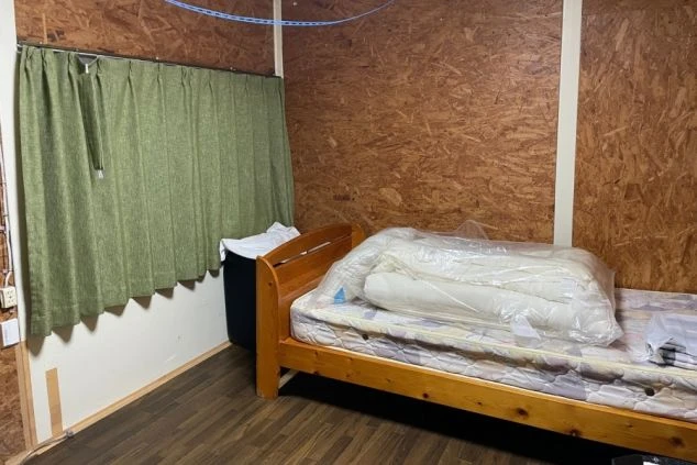 寮の居室。木製のベッドフレームとマットレス、サイドテーブルがあり、生活を始められる状態。