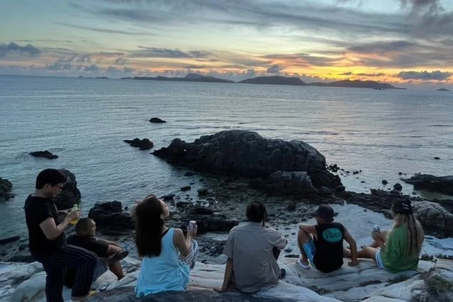 夕焼けに染まる海辺の岩場。友人たちが座って空を眺めており、ロマンチックな雰囲気が漂う。