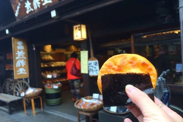 飛騨高山の古い町並みにある「手焼煎餅堂」と、手に持った海苔付きの大きな手焼き煎餅。食べ歩きグルメを楽しむ様子。