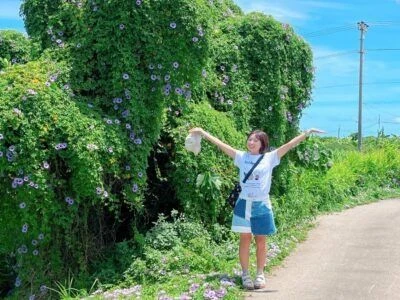 緑豊かな木々の前に立ち、両手を上げてポーズをとる女性。白いTシャツとショートパンツを着用し、楽しそうな表情をしている。背景には青い空と、木々の間から見える白い道が見える。
