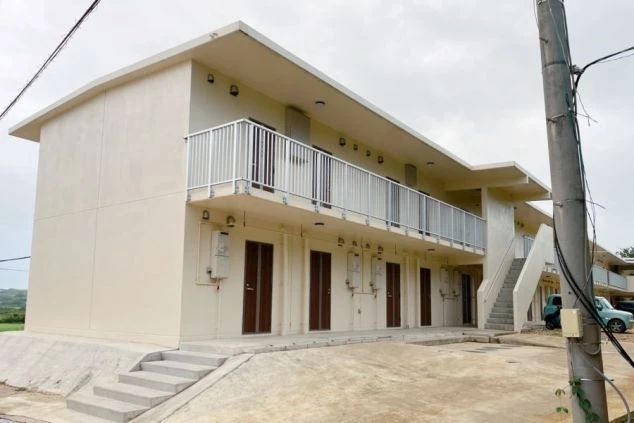 はいむるぶしの2階建ての寮アパートの外観。クリーム色の壁と、各階に並んだバルコニーが特徴的。新しい建物に見える。空は曇り空で、手前には電柱と電線が写っている。