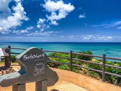 南国のリゾート地にある、海が見えるテラス席。木製の通路が海に向かって伸びており、通路の脇には緑の植物が茂っている。エメラルドグリーンの海と、青い空に白い雲が浮かぶ、美しい景色。