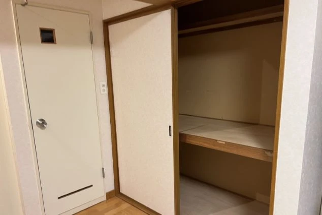 寮の個室の収納スペース。和風の引き戸を開けると、寝具などを収納できる広い中段付きの押し入れがあり、隣には白い扉の個室入口が見えます。