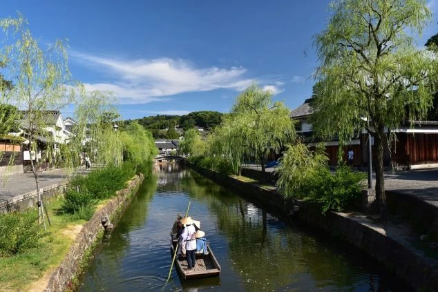 広角で捉えられた、晴れた日の川下りの様子。木々の緑が豊かな川の両岸には家々が立ち並び、川の中央には白い屋根の船が浮かんでいる。空は青く、白い雲が点々と浮かんでいる