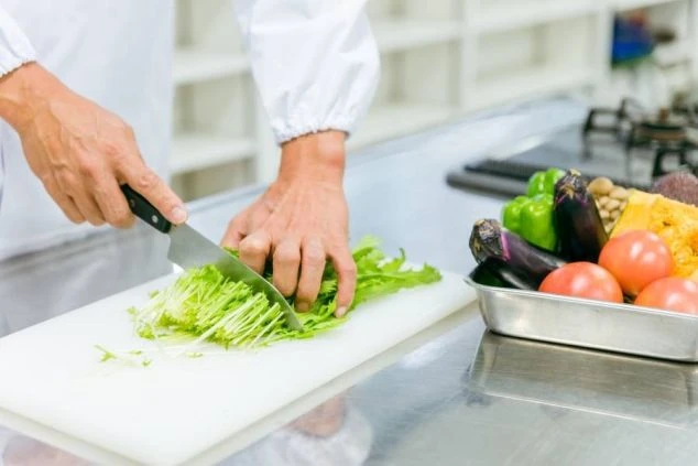  白い作業台でシェフが野菜を刻んでいる様子。シェフの手元には緑色の野菜があり、その右隣には色とりどりの食材が盛り付けられた皿がある。背景はぼやけている。