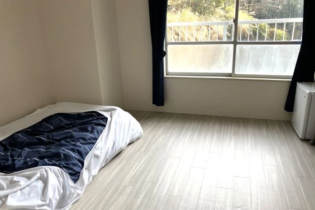 ワクトリの社員が出張で撮影した、リゾートバイトの寮の部屋の画像。フローリングの広々とした空間に、布団と小型冷蔵庫が置かれ、窓からは明るい日差しが差し込んでいる。