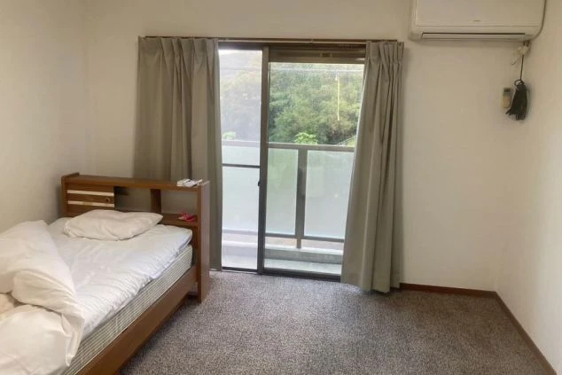 ベッドの足元から部屋全体を撮影した画像。部屋にはベッドとテレビ台があり、窓の外にはベランダと緑が見える。リゾートバイトの寮の部屋の画像。