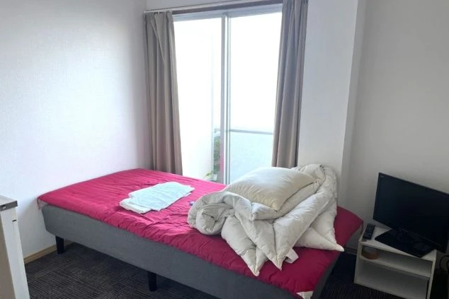 ベッドの足元から部屋全体を撮影した画像。部屋にはベッドとテレビ、そして窓がある。家具が少なく、広く使える。リゾートバイトの寮の部屋の画像。