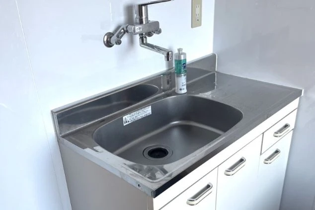 白い洗面台と、ステンレス製のシンクが写っている画像。シンクの下には収納スペースがあり、上には蛇口と洗剤が置かれている。リゾートバイトの寮のキッチンの画像。