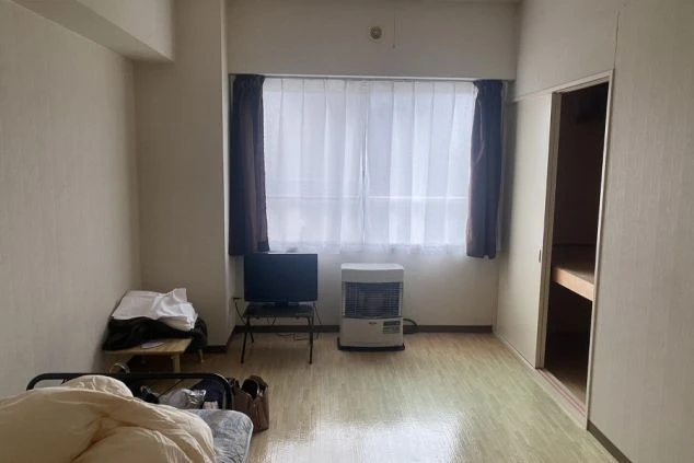 窓際にテレビとヒーターが設置された寮の個室。ベッドの横には収納力のある押し入れがあり、大きな窓からは自然光が差し込む。シンプルで機能的な一人暮らし向けの空間。