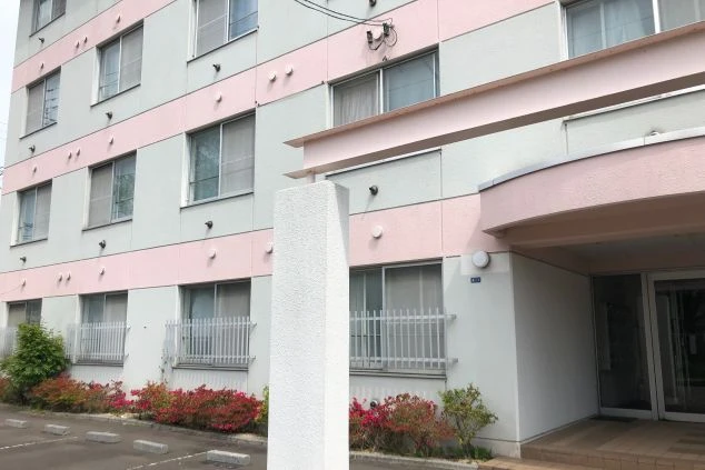 淡いピンクと白のラインが特徴的な、複数階建ての寮の外観。エントランス付近には赤い花が咲く植栽があり、手前にはアスファルトの駐車場が広がっている。