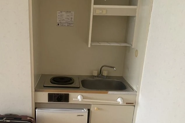 個室内に備え付けられたコンパクトなキッチン。1口の電磁調理器、小さなシンク、その下には小型冷蔵庫が収まっている。上部には棚があり、限られたスペースを有効活用している。