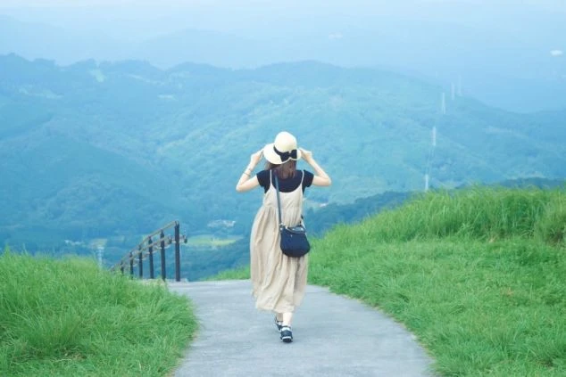 高原の道を歩く女性の後ろ姿の画像。麦わら帽子をかぶり、青い山々を背景に歩いている。リゾートバイトの休日に楽しめる開放的な風景。