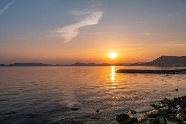 海に沈む夕日の絵。水面には夕日の反射が光の筋となり伸びています。地平線には陸地がシルエットになって見え、空には日が残っているため水面近くにオレンジと黄色の光が、上空には青みがかった色が広がっています。