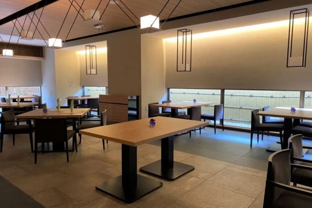 モダンで和風なデザインのレストラン。木製テーブルと椅子が並び、窓の外には竹垣のような目隠しが見える。長方形のペンダントライトと間接照明が空間を照らしている。