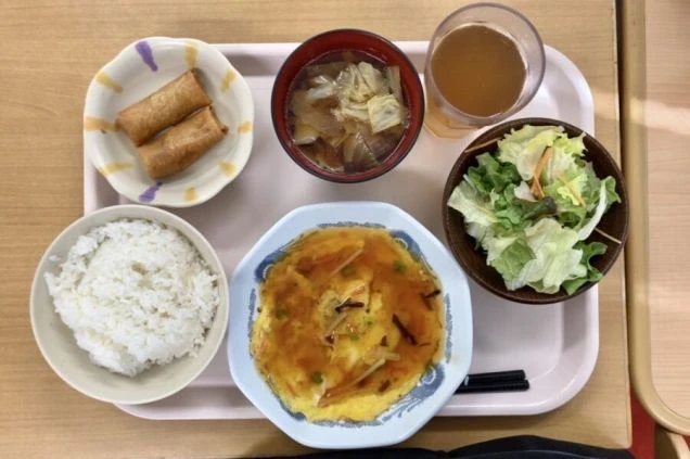 従業員食堂または寮のまかないと思われる食事。トレイの上に、天津飯（かに玉）、ご飯、春巻き、味噌汁、サラダ、飲み物（お茶または麦茶）が並んでいる。