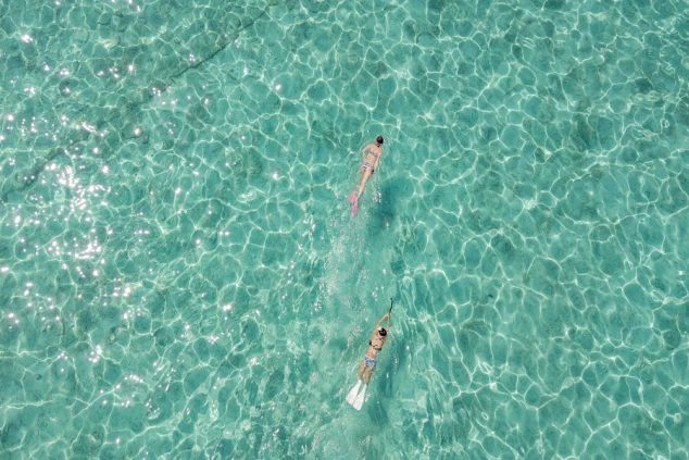 非常に透明度の高いターコイズブルーの海を上空から捉えた写真。水面に光が反射してきらめく中、水着を着てシュノーケルやフィンをつけた2人の人物が水面を泳いでいる。